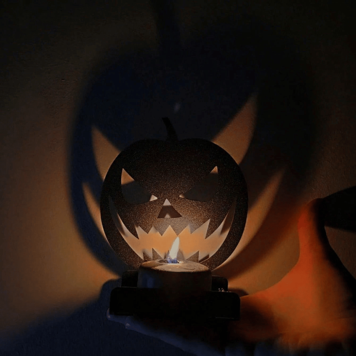 Black iron jack-o'-lantern tealight holder lit, casting a large flickering pumpkin face shadow on the wall